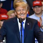 Man in suit speaking at rally with red hats.